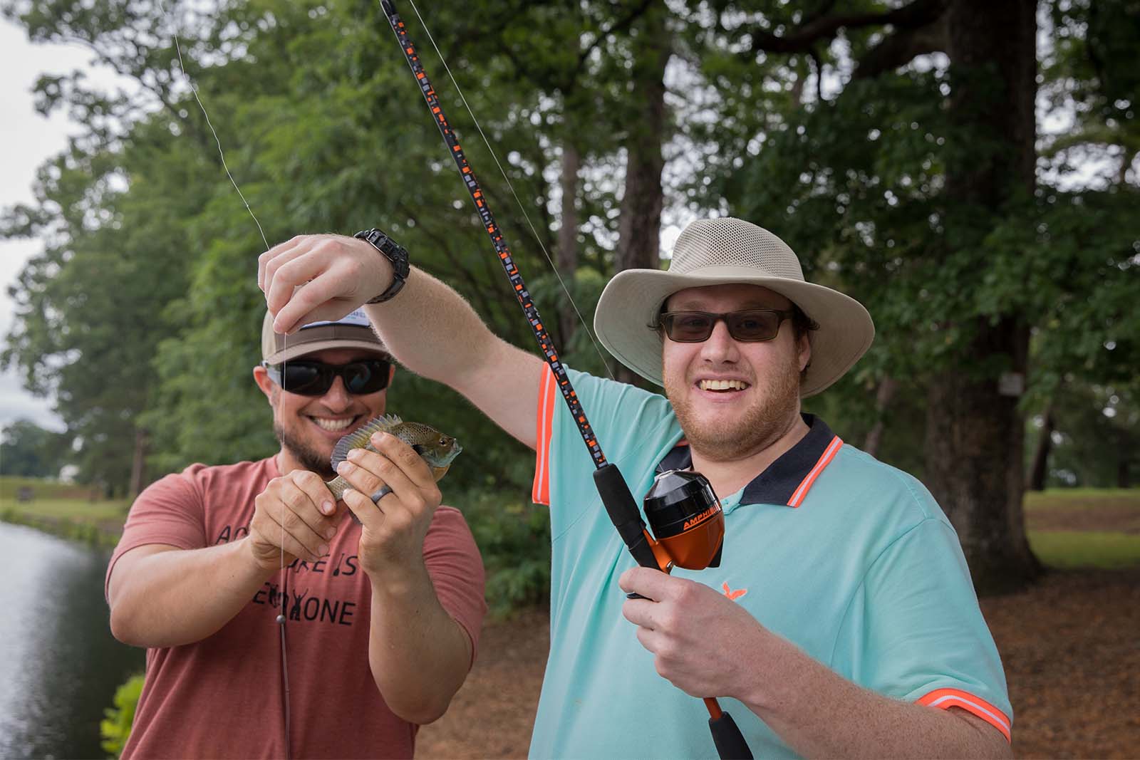 两个男人站在湖岸边。一个人拿着鱼竿，另一个人拿着一条从鱼钩上取下来的鱼