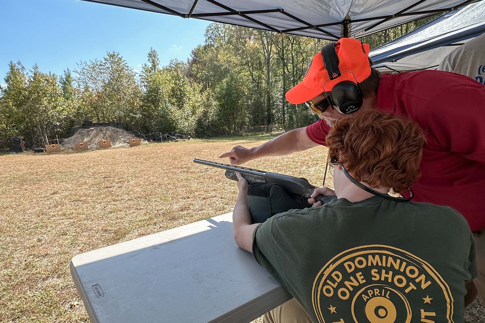 一名男子正在练习用猎枪射击目标；一名教练站在旁边指向目标