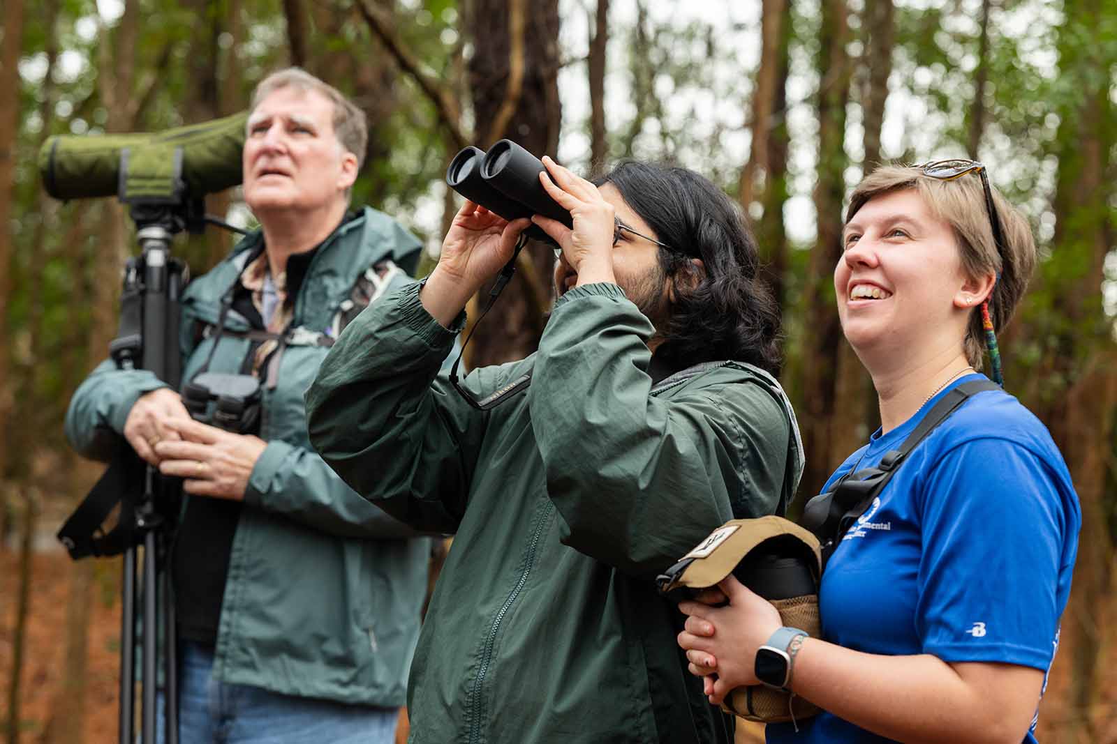 两个穿着深绿色夹克的人向上看，其中一个人用双筒望远镜