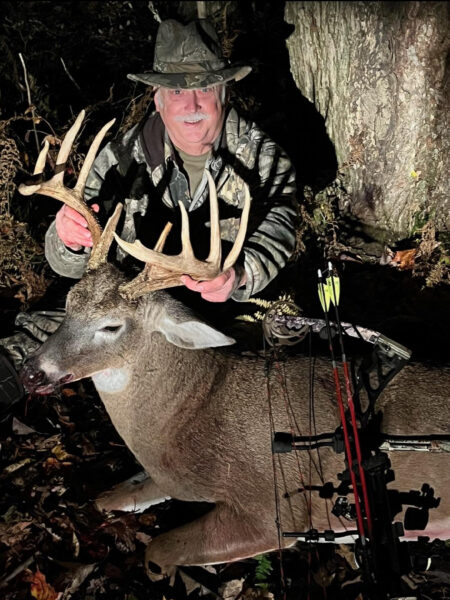 照片中，一名身着迷彩服的男子手持弓箭，手中握着一只捕获的大雄鹿的鹿角。