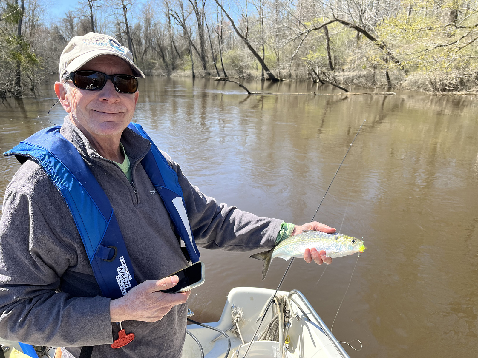 一名男子身穿救生衣，站在河中的船上，手里拿着一条闪闪发光的鲱鱼。
