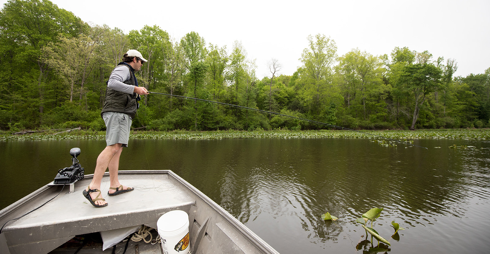 一名男子站在平底船的船头，手握鱼竿，望着水面。
