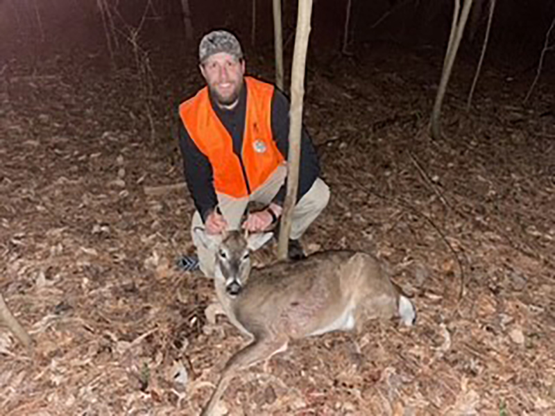 照片中，一名身穿橙色背心的男子跪在地上一只小白尾鹿后面。