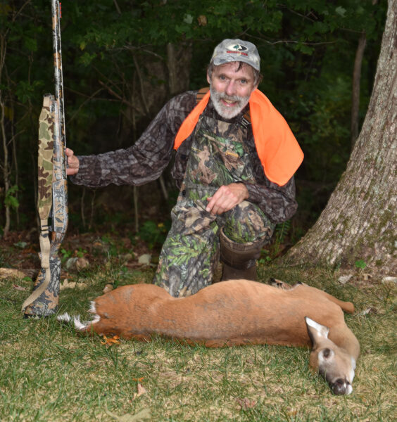 照片中，一名身穿橙色迷彩服的男子手持枪支，跪在地上一只白尾鹿的后面。