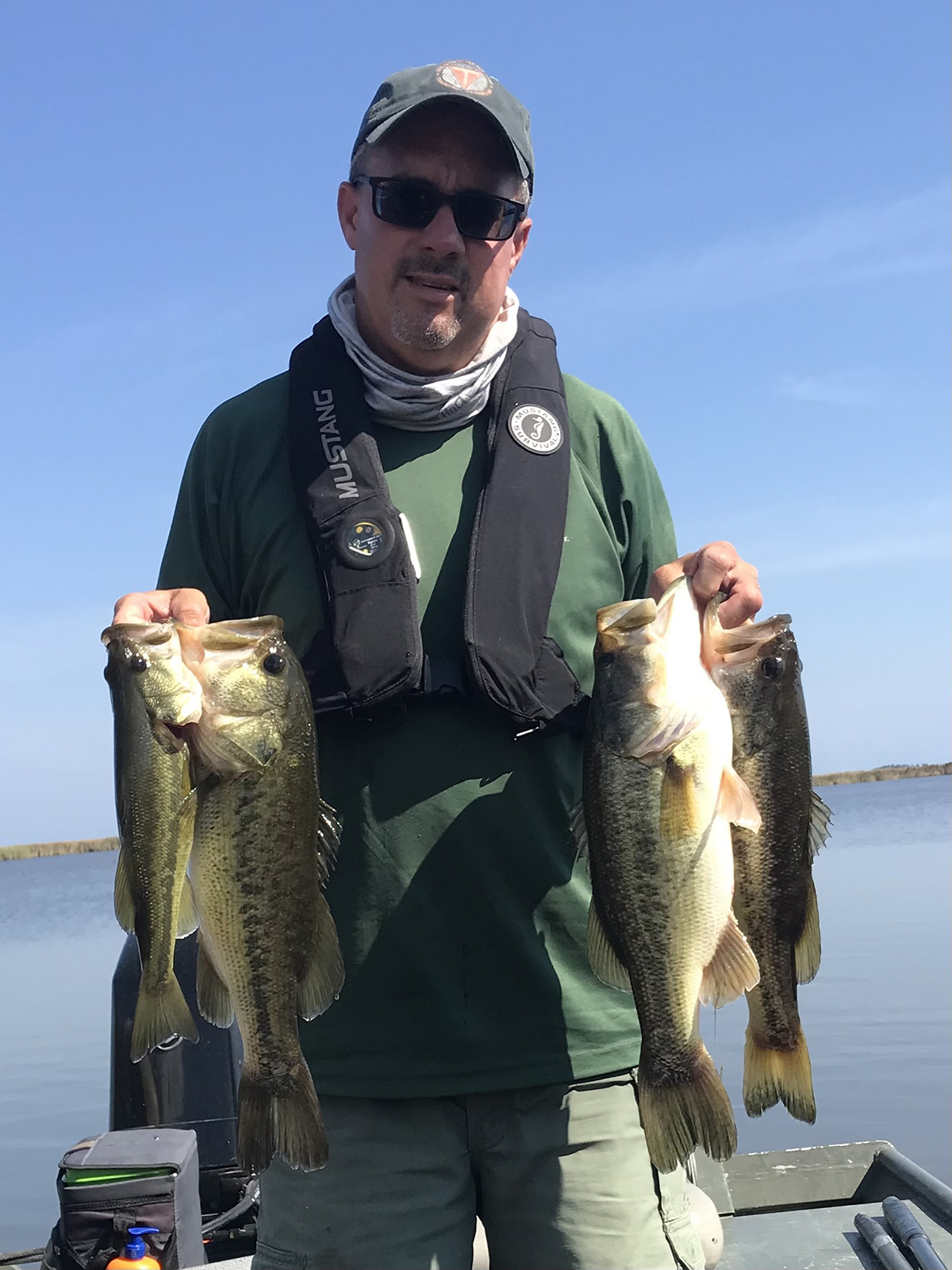 一名男子查德·博伊斯 (Chad Boyce) 站在一艘渔船上，手里抓着四条大嘴鲈鱼。他身穿绿色上衣、救生衣，戴着太阳镜和棒球帽。