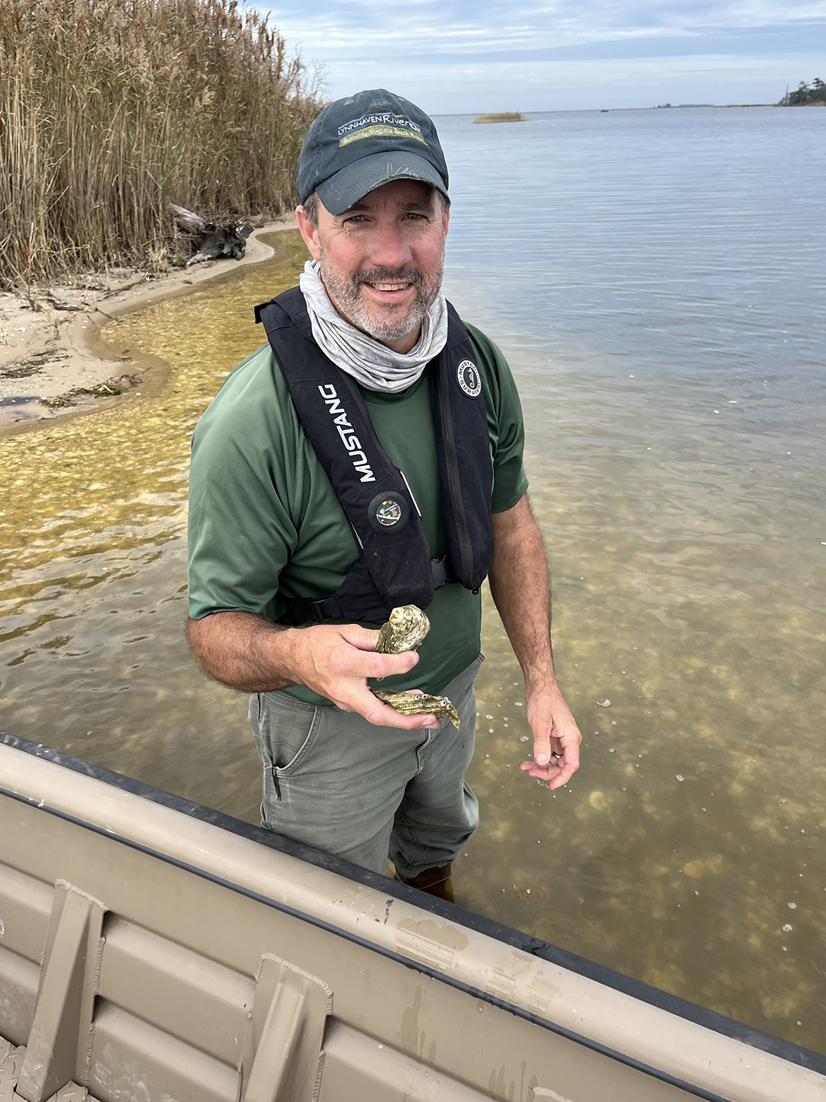 照片中，一名男子站在船旁齐膝深的水中，手里拿着牡蛎壳。 