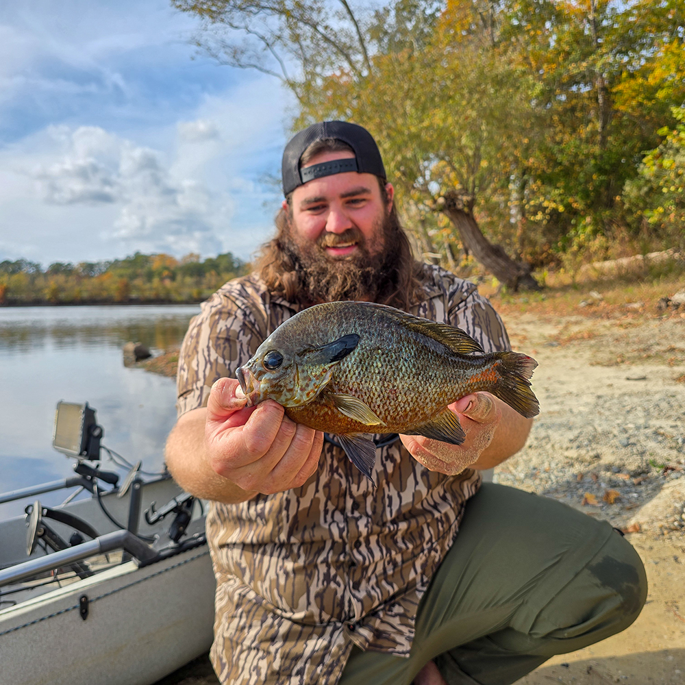 照片中，一名男子跪在岸边的一艘皮艇前，手里举着一条大红胸太阳鱼，面带微笑。