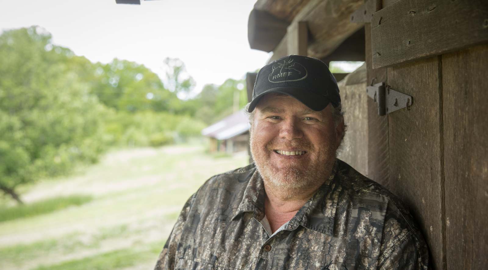 一张留着胡须的男子站在一座粗糙的木质建筑外的肖像照片。