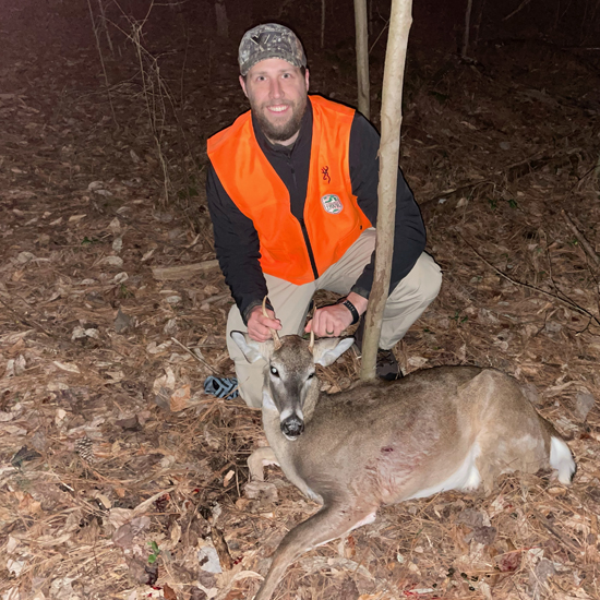 照片中，一名身着橘红色衣服的男子跪在地上，面前躺着一只尖头野鹿、